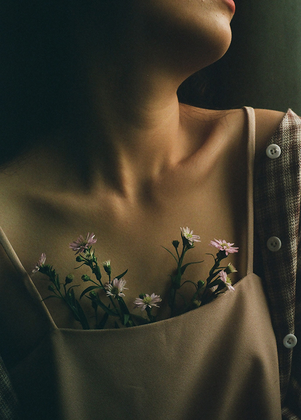 Warm but darkly-lit photograph of a woman's chest. She is wearing a camisole and overshirt. Flowers peak out from the top of the blouse.