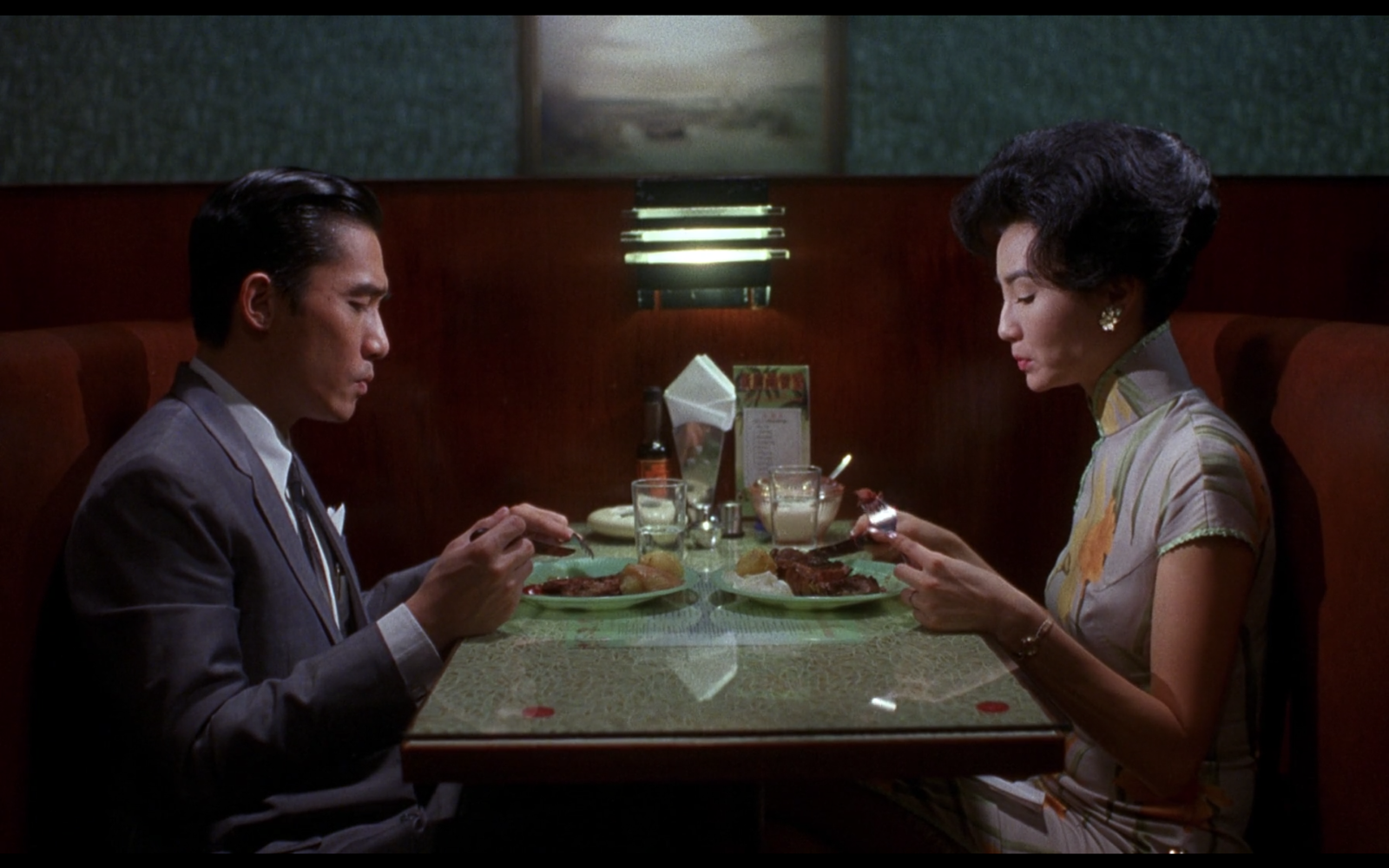 A screen still from the film In the Mood for Love, featuring the two main characters, Mrs. Chan and Chow Mo-wan, sitting across from each other as they eat in a restaurant.