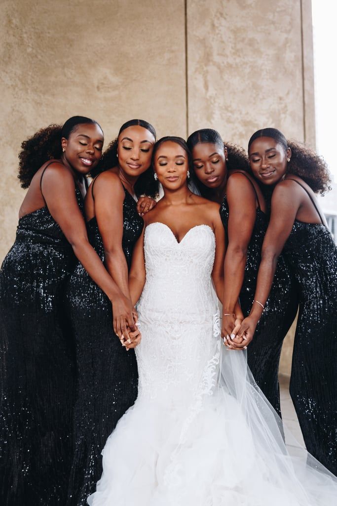 Noiva, com vestido branco rendado tomara que caia, ela é uma mulher negra e usa o cabelo preso num coque. Suas madrinhas, duas de cada lado da noiva, estão com a cabeça encostada nela e seguram suas mãos. 