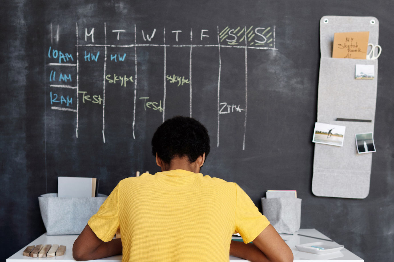 A young man sitting on his desk focused on studying while facing a weekly plan written on a black board on the wall.