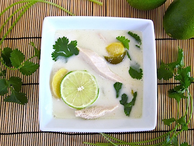 Overhead view of a bowl of Chicken Coconut Soup on a bamboo mat with cilantro and lime on the side