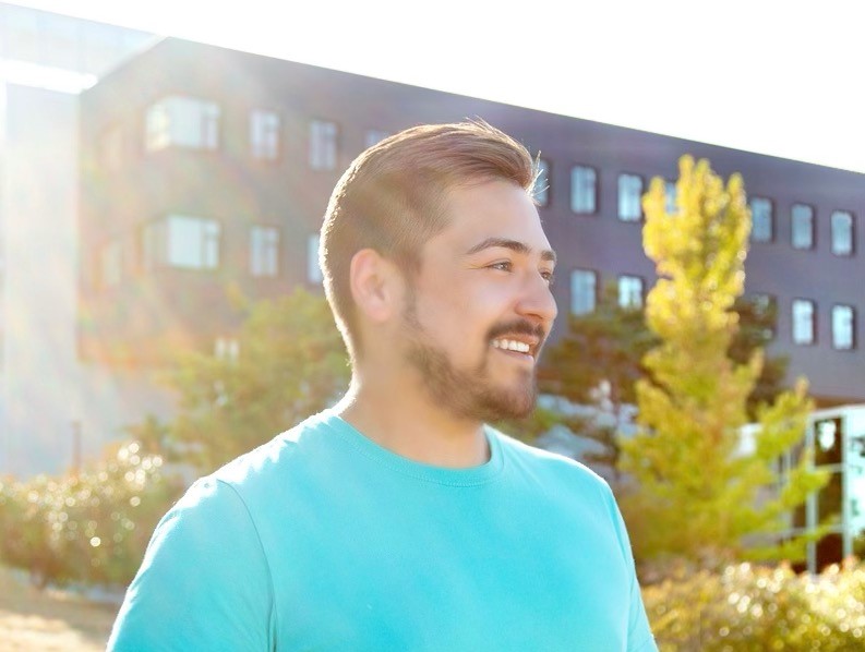 Young man in blue t-shirt smiles in front of a building on a university campus.
