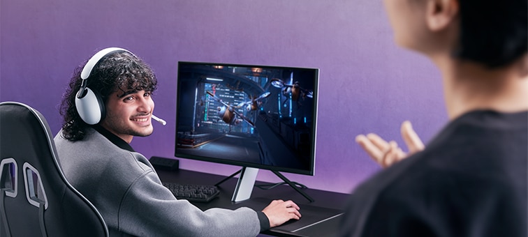 Man wearing INZONE H9 headset sitting at a desk with monitor and game on screen, talking to partner in foreground right