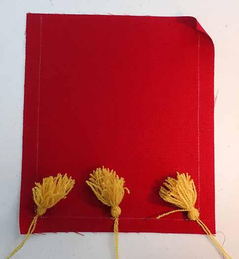 A wide shot of a piece of red wool fabric with three golden yellow tassels pinned at the bottom edge. The left, bottom, and right edges have a thin white line marking a 1/2" seam allowance.