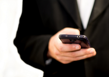 A close-up view of a person holding a black smart phone. The person appears to be wearing a suit. 