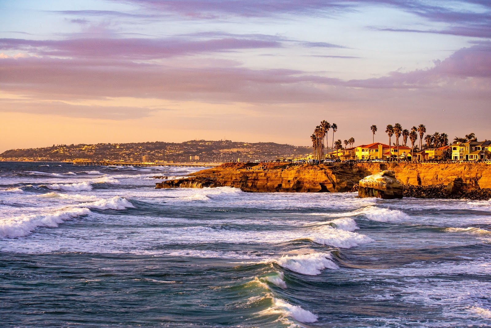 blue ocean waves coming to san diego shore with short cliff and bright buildings and palm trees during sunset in california