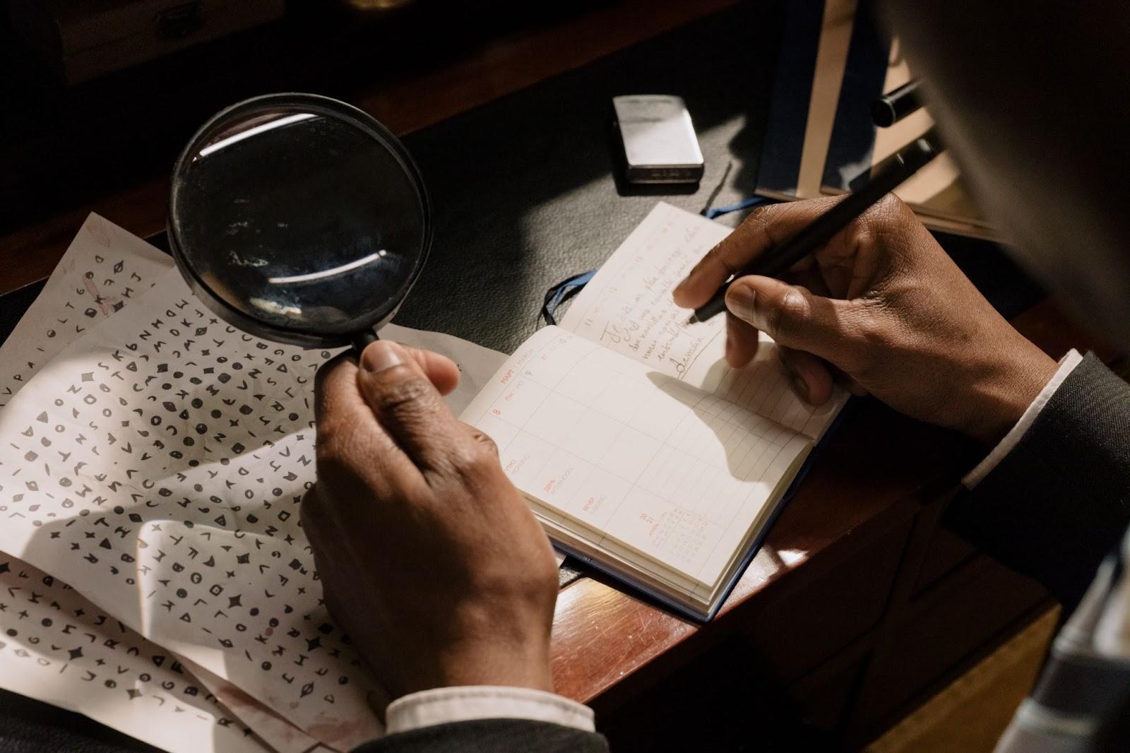 Man Holding Magnifing Glass and Writing in Small Journal