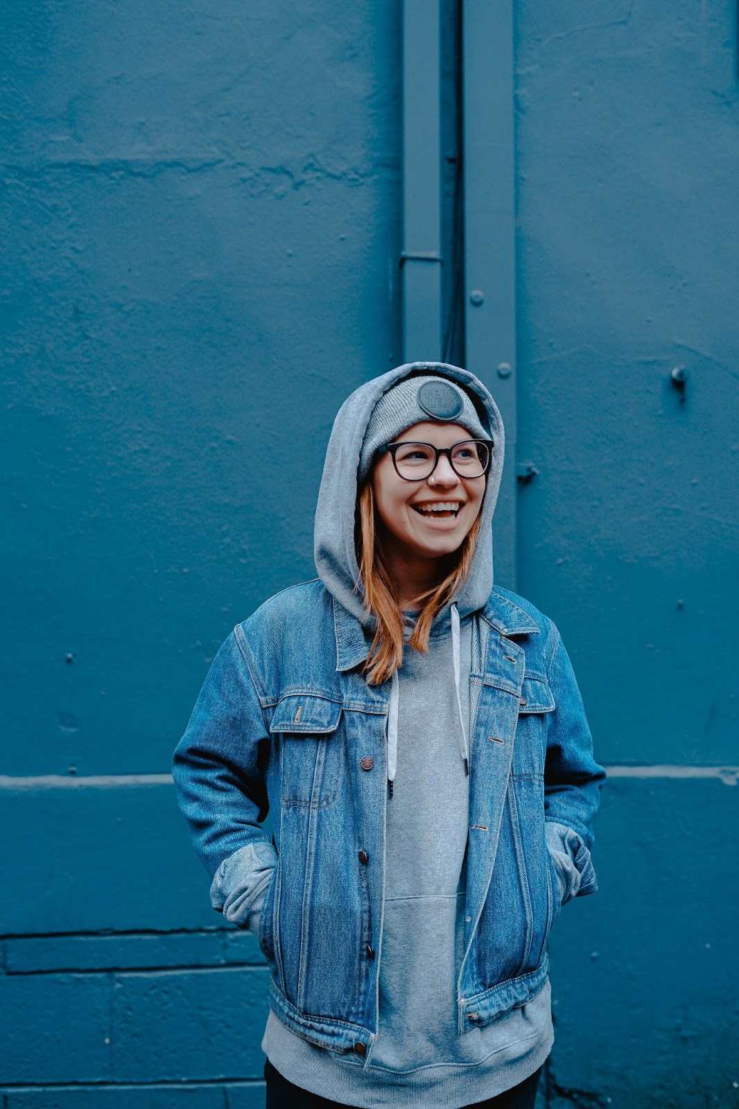 A picture of a smiling teenage girl in glasses, wearing a jean jacket and gray hoodie against a blue background.