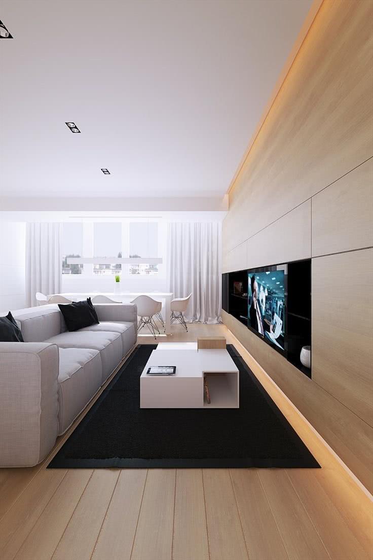 Sala com painel de TV de madeira ocupando toda parede, sofá cinza com almofadas pretas, piso de madeira, mesinha de centro branca e tapete preto.