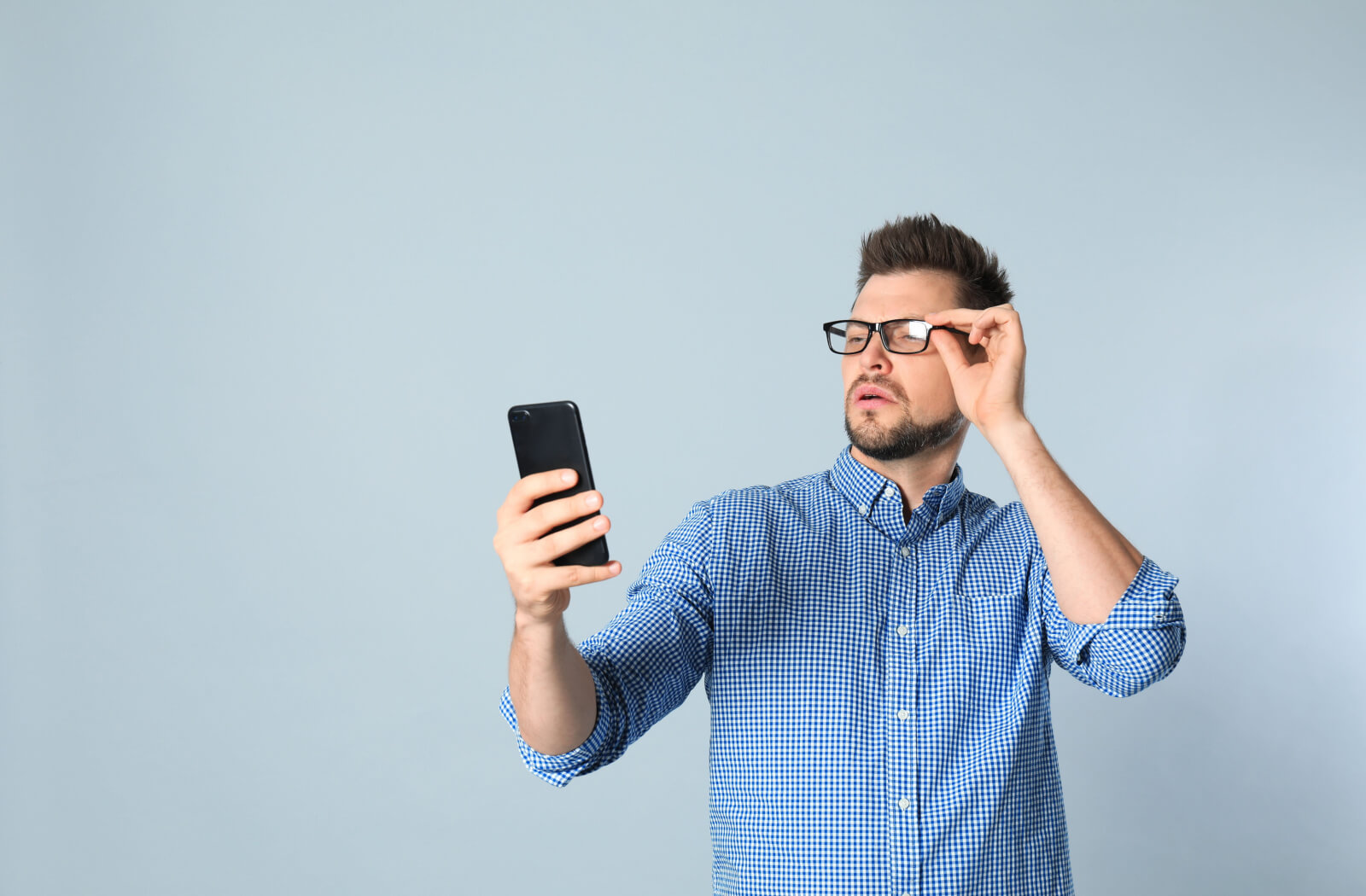 A man wearing eyeglasses is squinting while looking at his smartphone while holding it a bit far from him