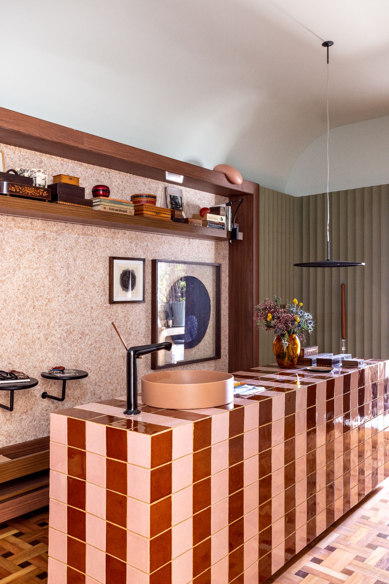 Revestimentos Casacor: Ambiente com piso de tacos de madeira, parede com revestimento de granilite rose, prateleiras de madeiras com objetos e bancada com revestimento marrom e rose.