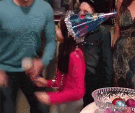 GIF de uma menina comemorando, em meio a outras pessoas. Ela usa um chapéu de festa de aniversário, se empolga na comemoração e acaba jogando um vaso de vidro que está na mesa, no chão. 