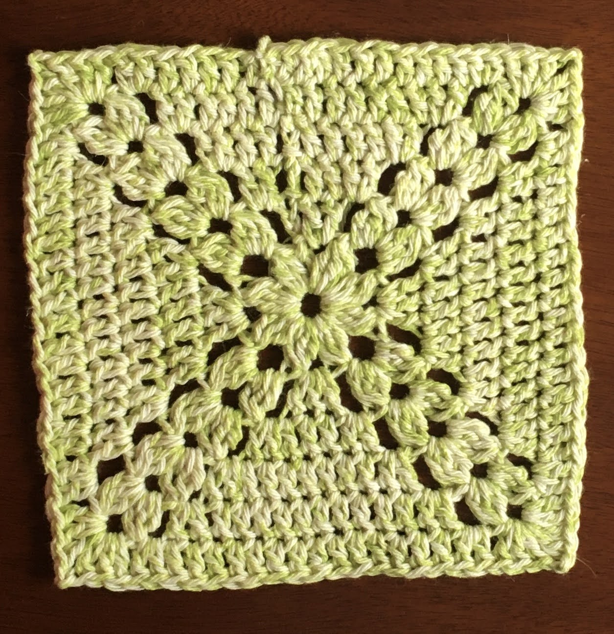 A crocheted green square, with stitches that create a thick X-shaped pattern in the center.
