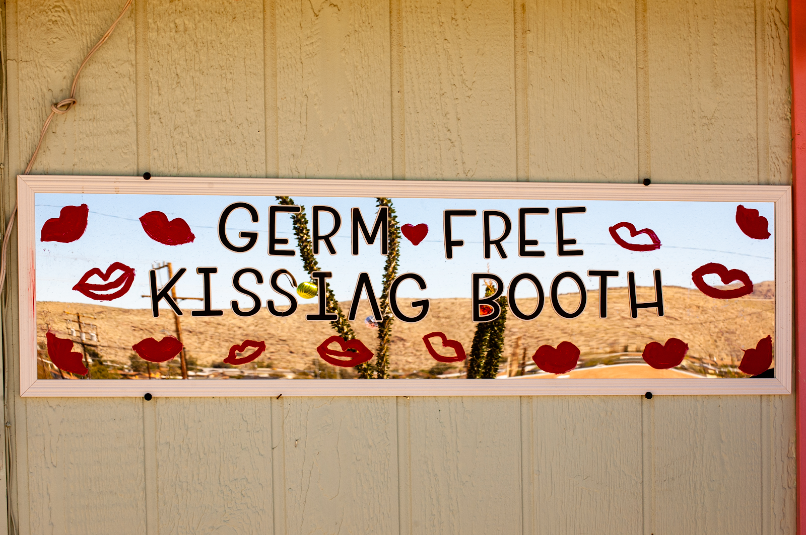 A mirror, laid sideways, and painted with a series of red lips, says "Germ Free Kissing Booth" in black letter across it. It is mounted on an exterior wall of the Beauty Bubble hair salon, Joshua Tree, California. April 24, 2020. (Julie Dole/Pirate)