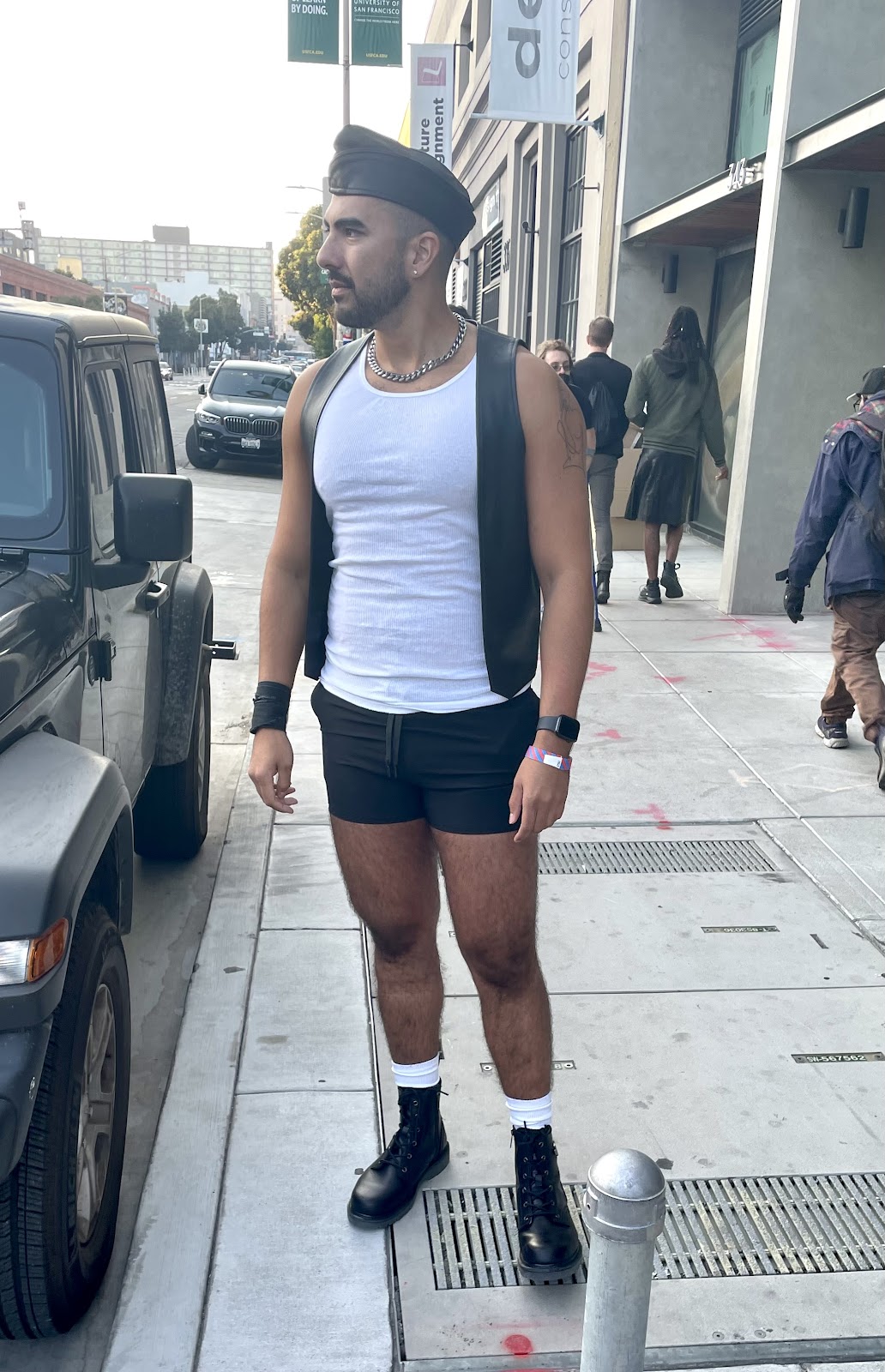 Gay male wearing a white ribbed tank top and a gay leather vest posing on the sidewalk outside of Folsom Street fair 2023 in San Francisco