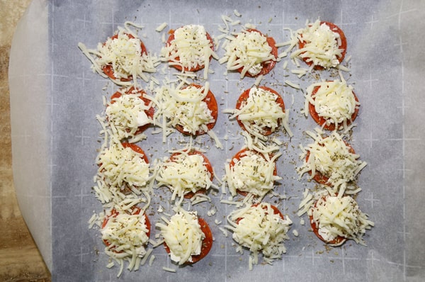 Pepperoni slices sprinkled with grated cheese and seasonings arranged on a baking sheet with parchment paper