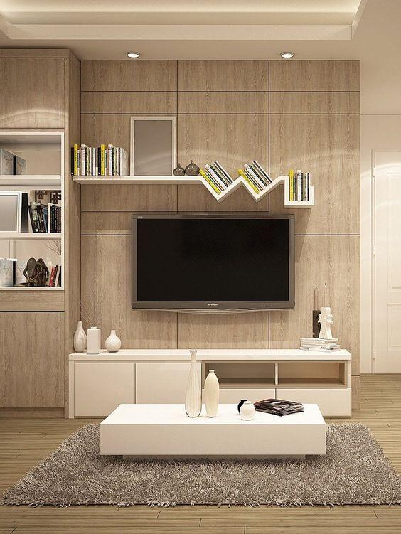 Sala de TV com móvel planejado de madeira clara reproduzindo painel de TV, prateleiras decorativas com livros, rack branca, mesinha de centro branca, tapete cinza e piso de madeira. 