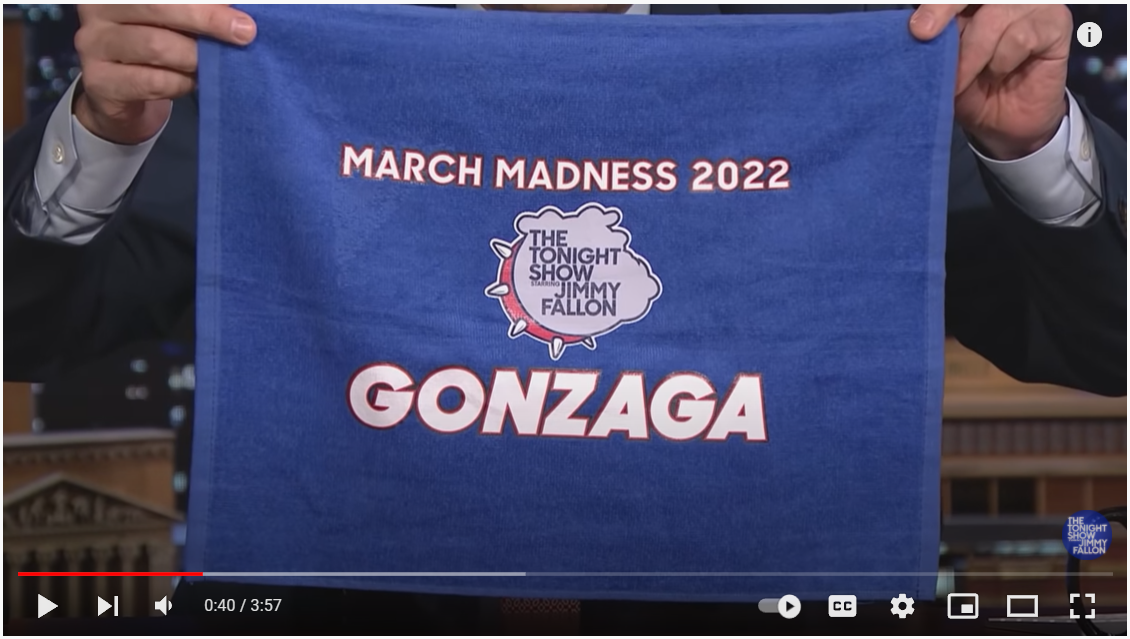 Jimmy Fallon holding up a blue towel with "March Madness 2022" and "Gonzaga" on it, with the Tonight Show logo in a bulldog head.