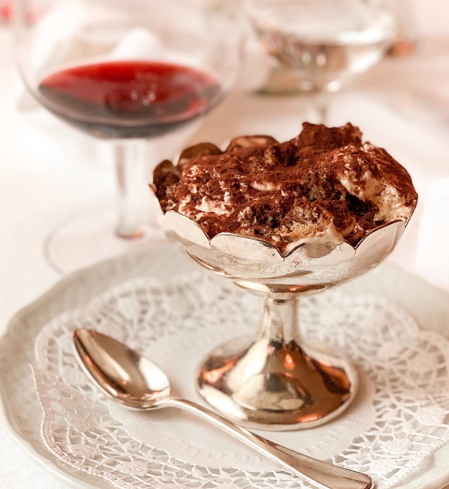 Tiramisu in a silver bowl with a spoon and glass of red wine in the background.