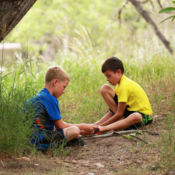 Two young boys participating in summer camp through CSU’s Environmental Learning Center sit in the grass beneath a tree, talking and observing something on the ground.