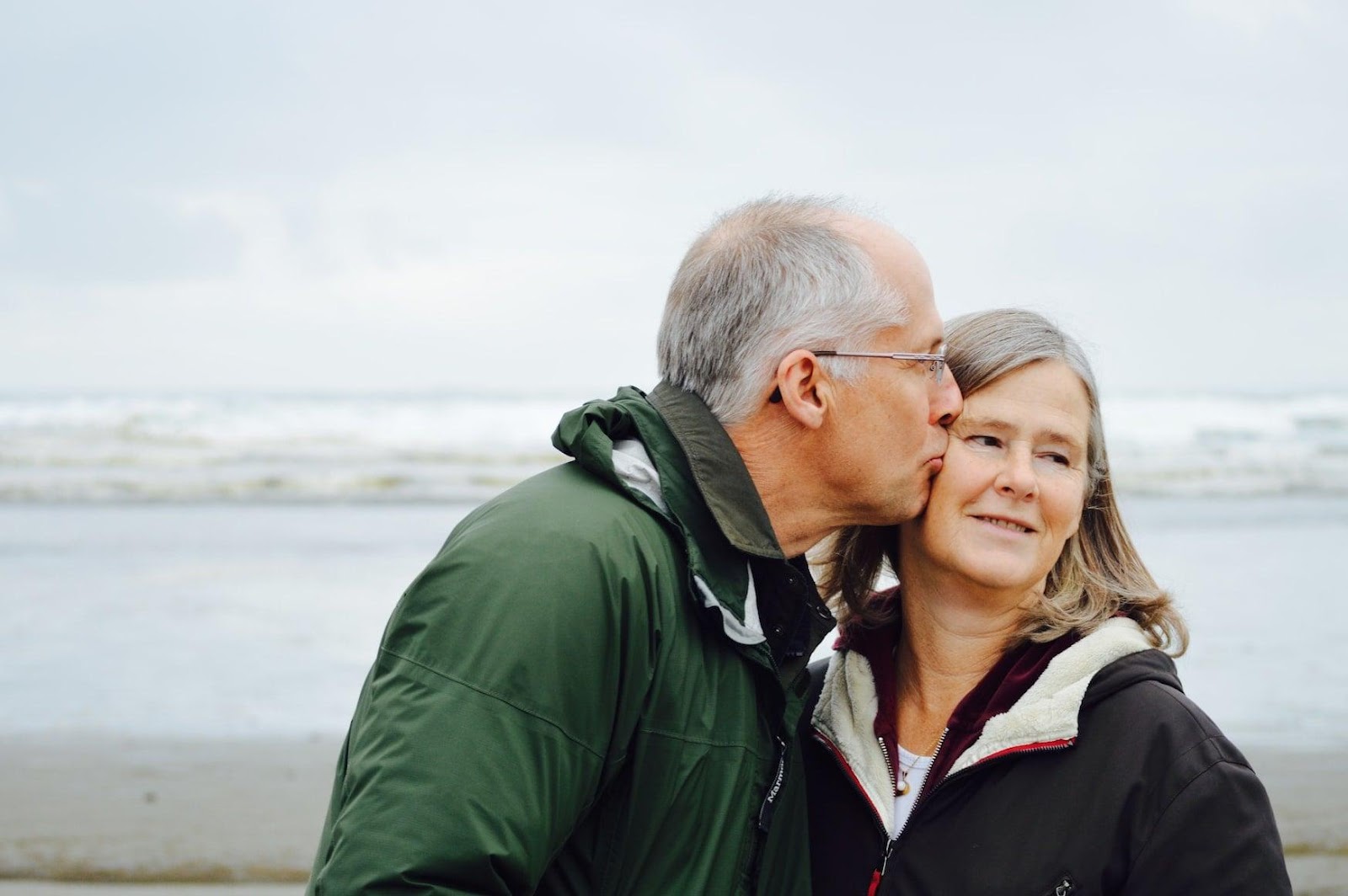 An old man kissing an old woman on the cheek with the beach on the background 