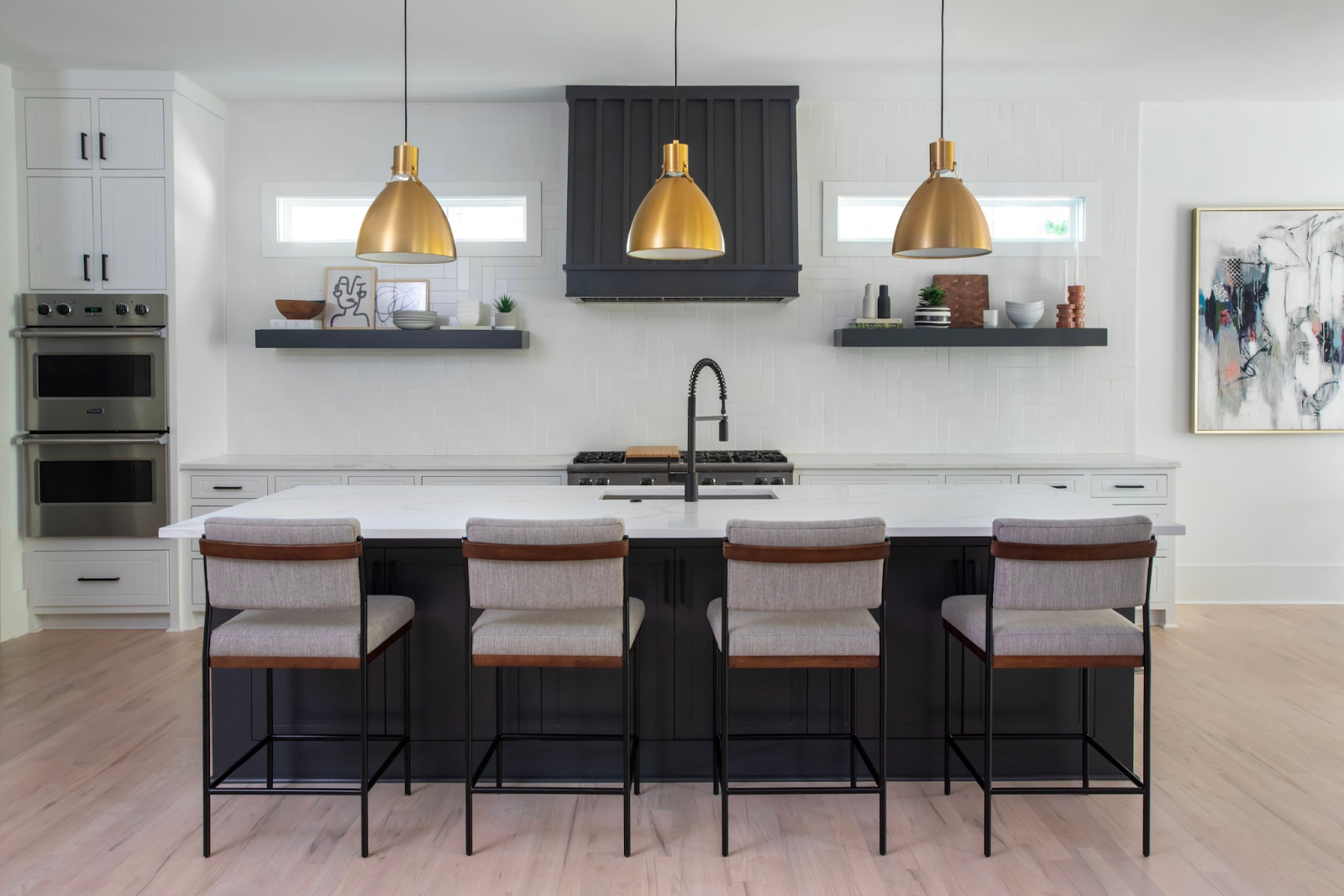 Three gold metal kitchen island pendants hanging over a white kitchen island with 4 gray and brown chairs. In background is ceramics and art sitting on shelves, a black sink and faucet, and black stove. 