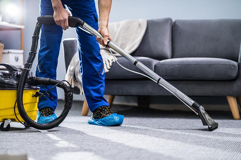 WeServe | A person deep cleaning the grey carpet with a yellow vacuum while wearing blue protective bags on his feet with a grey couch in the background