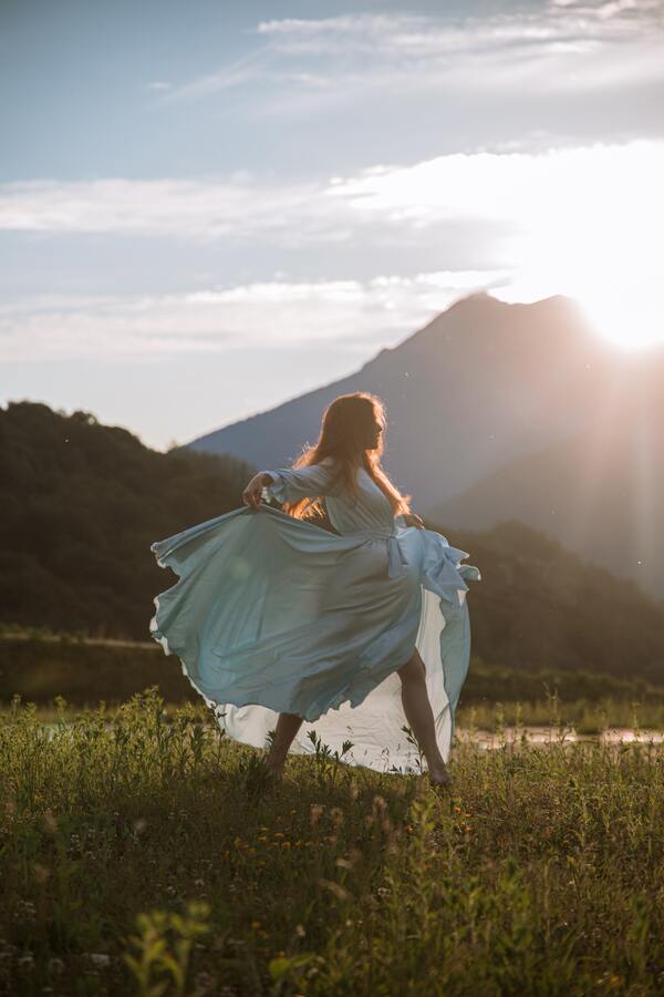 Foto de uma mulher em um campo aberto com um vestido azul claro longo, dançando sob o Sol. 