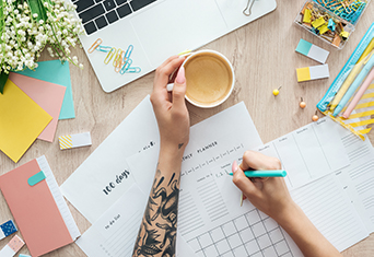 Two hands - one with a tattoo holding a cup of coffee and the other one a turquoise pen. She's on a desk with office stationery and a laptop.