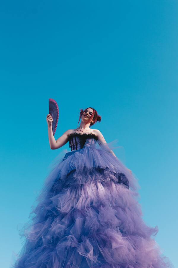 Foto tirada debaixo de uma mulher com um vestido roxo cheio de babado segurando um leque.