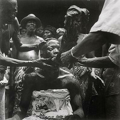 Reprodução fotográfica do artista. Imagem representativa de um culto de iniciação a religião de matriz africana. O personagem negro em foco, está sentado com olhos fechados, seu corpo está envolvido por um pano branco. Um homem à sua esquerda, segura sua cabeça, enquanto o outro posicionado à sua direita, segura uma galinha preta na direção de sua boca. É possível ver manchas de sangue.