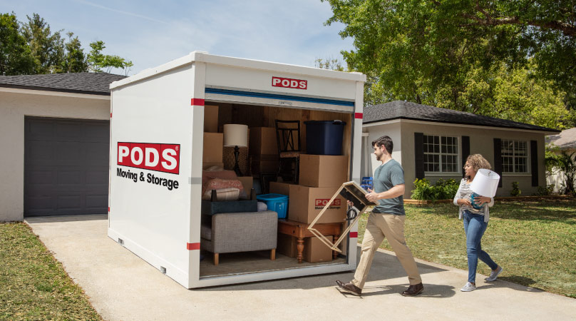 A man and woman are loading more furniture into their mostly loaded PODS moving and storage container in their driveway. The man is carrying a side table, and the woman is carrying a lamp.