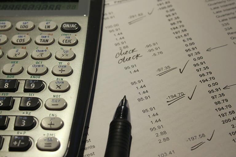 A calculator and pen next to a spreadsheet marked up with financial tallies.