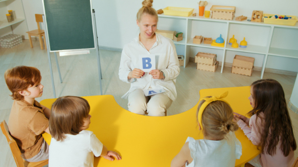 A teacher holding a letter flashcard and teaching four children seated at a round, semicircular, yellow table.