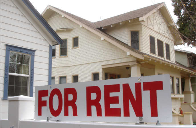 A close-up of a “FOR RENT” sign in front of a residential home with a neighboring home in the background