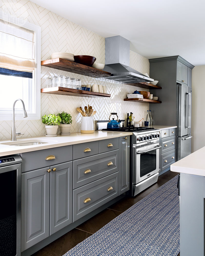 modern galley kitchen with grey cabinets, white herringbone backsplash, brass cabinet hardware, wooden open shelves and stainless steel appliances