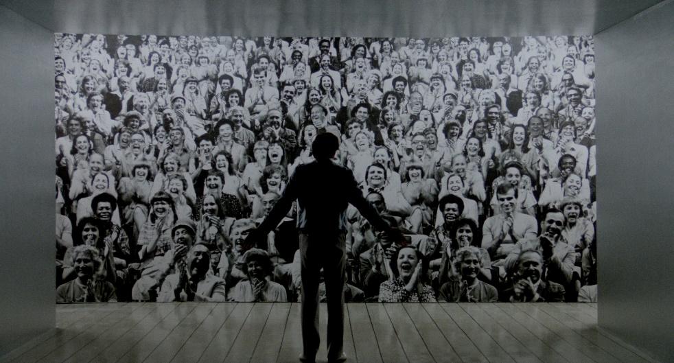 A screen still from the film The King of Comedy, featuring Robert De Niro as Rupert Pupkin, standing in front of and facing a wall covered in a print of a smiling and laughing audience.