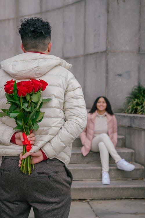 Back view of anonymous guy with curly hair in warm jacket holding bouquet of fresh red roses while standing in front of smiling ethnic girlfriend sitting on stairs in park