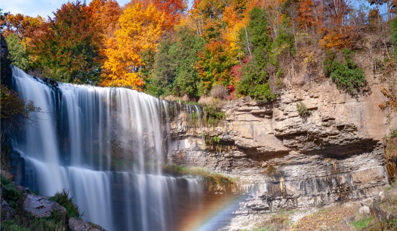 A Niagara Escarpment waterfall in Hamilton, Ontario, creates a rainbow in the mist. The leaves of the surrounding trees show bright yellows and oranges as they change colour for the fall.