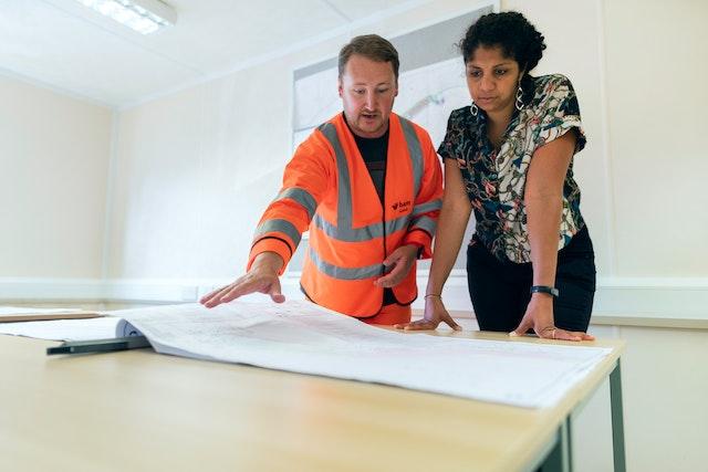 A man in work overalls explaining something to a woman in front of a blueprint spread on the table as taking professional advice is very important if you want to save money on your home renovation.