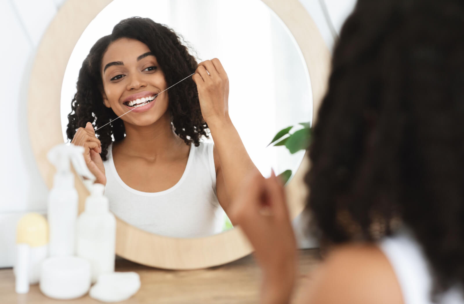 A young woman in the mirror holding dental floss on her hands and flossing her teeth.