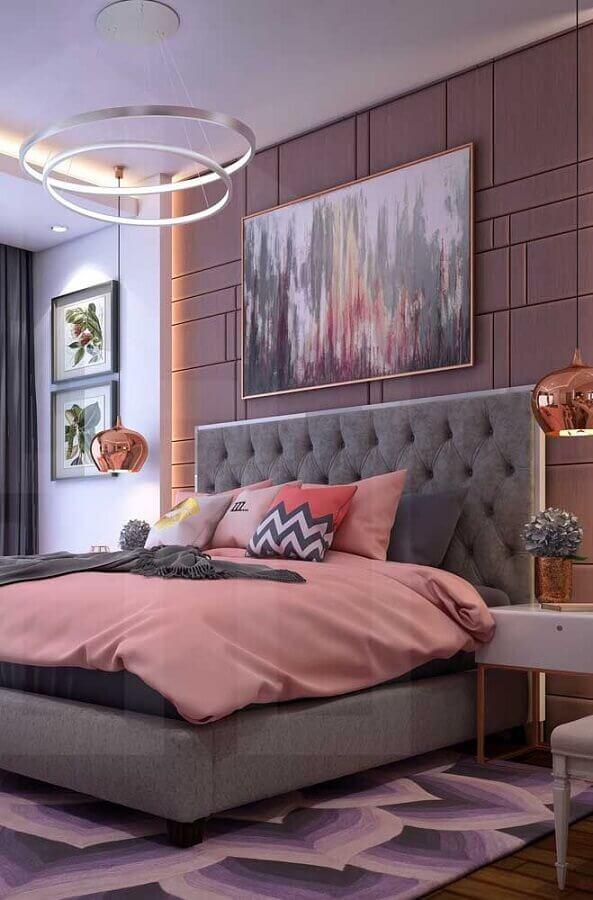 Quarto de casal com decoração em tons de rosa e cinza, cabeceira da cama estofada cinza, parede de fundo rose com molduras, móveis brancos, piso de madeira com tapete rosa e cinza e luminária pendente prata