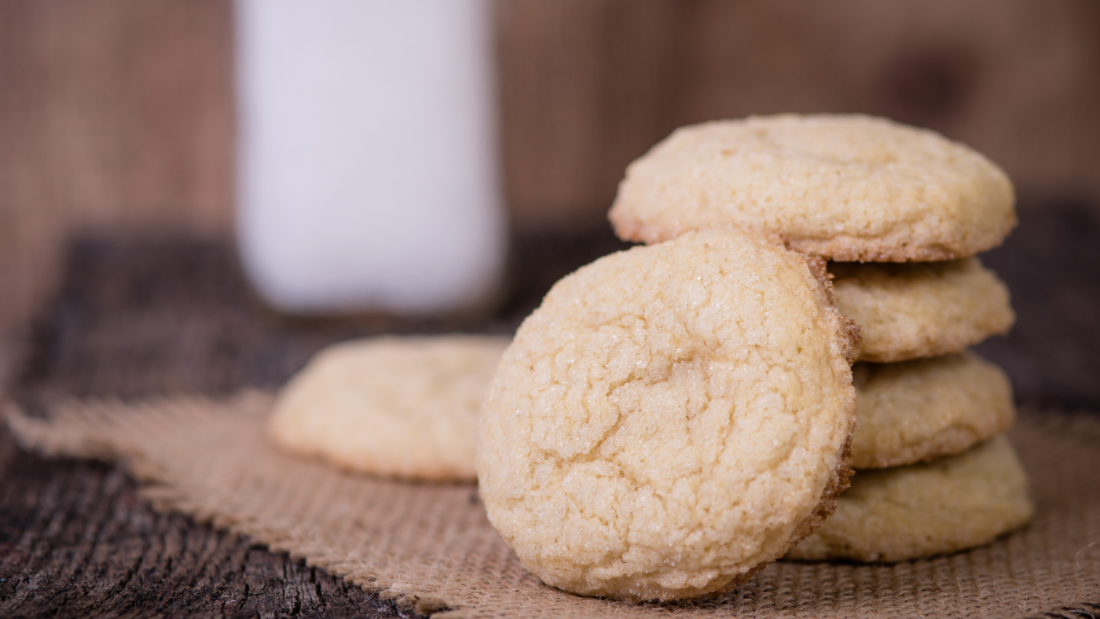 four .sugar cookies stacked with one leaning against the stack in the front, one laying behind the stack to the left, and a glass of milk in background