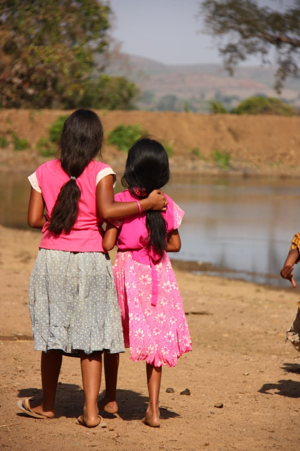 Two young girls are walking side by side. You can see them from the back. One of them has their arm around the other one's back. It is a village landscape with a lake and some greenery as well as mountains in the background.