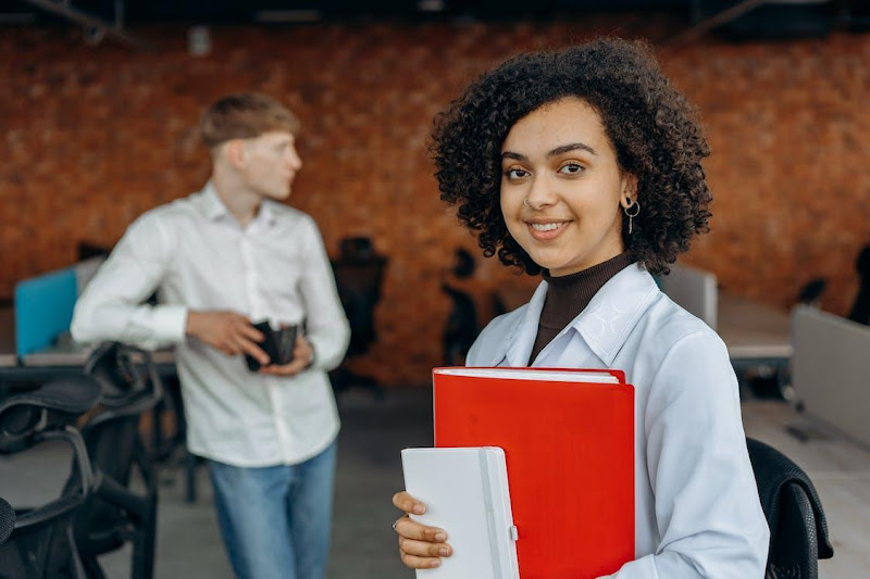 Free Young Woman in White Long Sleeve Shirt Holding Financial Book Report and Standing with Her Colleagues Inside an Office    Stock Photo