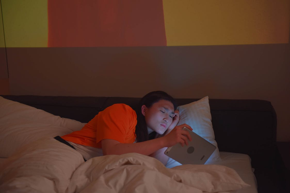 man in orange shirt laying in bed while holding tablet
