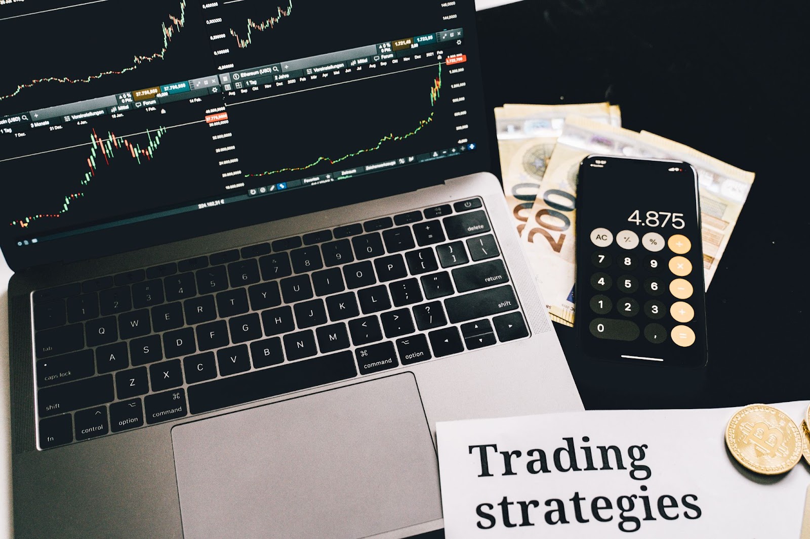 Stock charts on a laptop, calculator on a phone, and a trading strategies paper