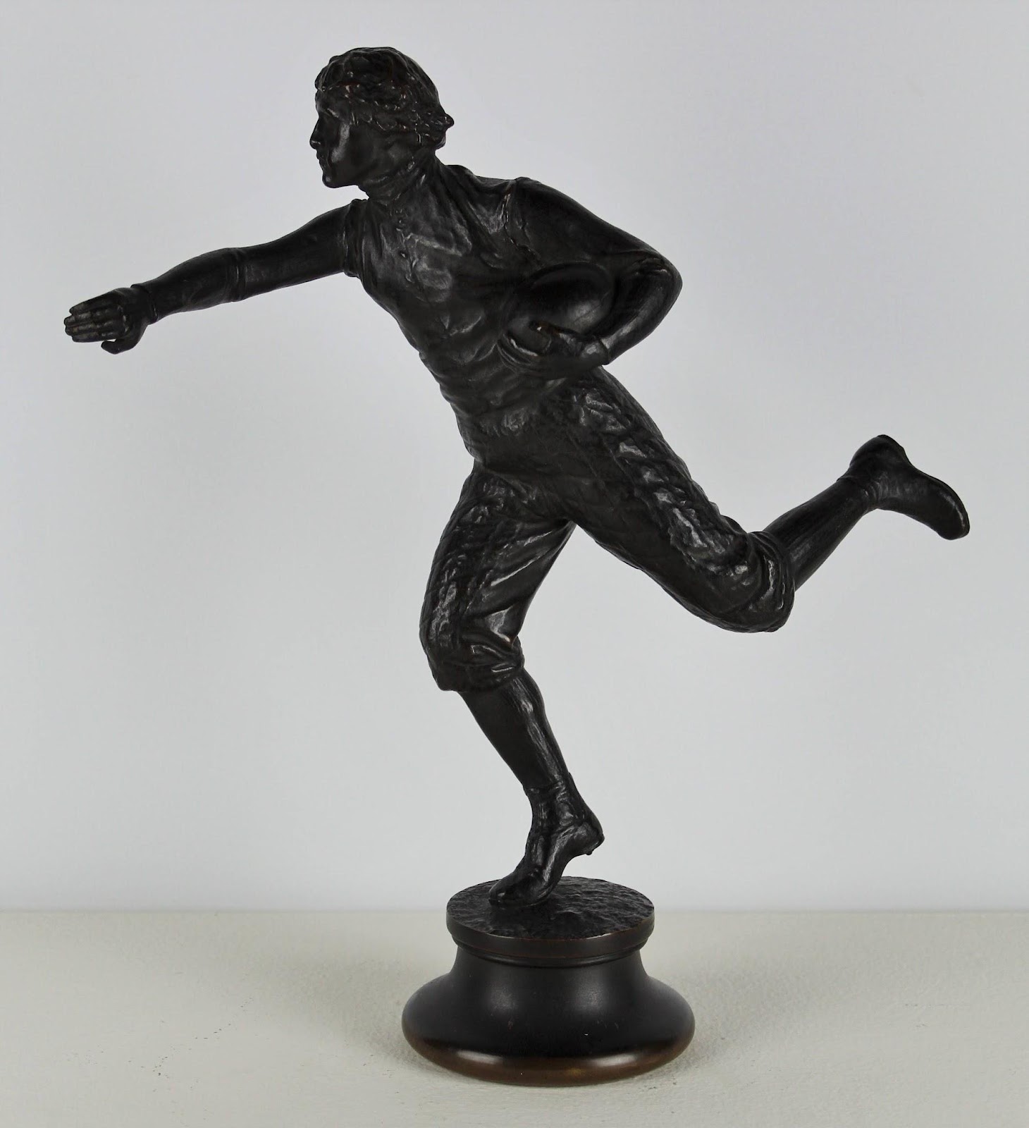 A bronze sculpture of a football player, one arm cradling the football to his chest while the other is extended to push away an invisible opponent. He stands balanced on on the toe, his other leg extended behind him in a run.