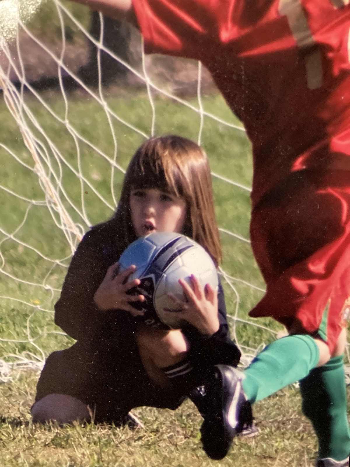 A girl (Kyla as a child) kneels down and catches a soccer ball. An opposing forward in a red jersey, green socks and black cleats is to the right.