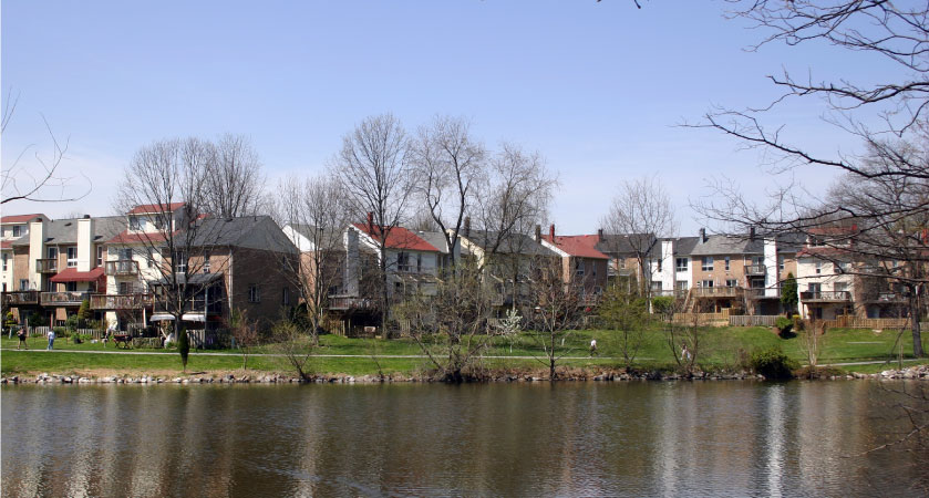 The back view of residential buildings that share a large greenspace and a lake with a walking trail in Columbia, Maryland.